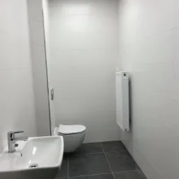 Nowoczesna, minimalistyczna toaleta z białymi płytkami ściennymi, szarą podłogą z płytek, wiszącą miską WC i umywalką z chromowaną baterią, oświetlona g&oacute;rnym światłem.