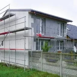 Dom jednorodzinny w trakcie prac, elewacja częściowo pokryta szarym materiałem izolacyjnym, widoczne rusztowania i betonowe ogrodzenie.