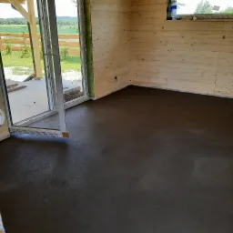 Świeżo wylana, ciemnoszara posadzka w pomieszczeniu z drewnianymi ścianami i otwartym oknem balkonowym z widokiem na zewnątrz.