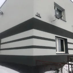 Nowoczesna elewacja domu dwukondygnacyjnego z poziomymi pasami w kolorze szarym i białym, widoczne dwie anteny satelitarne i tabliczka z numerem 94 na tle zimowego krajobrazu.