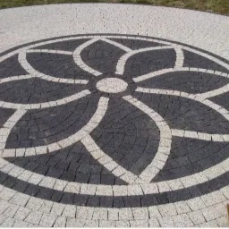 Okrągły podjazd z kostki brukowej w dwóch kolorach, ułożony we wzór kwiatowy, z widocznym ubijakiem w prawym dolnym rogu.