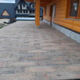 Szeroki taras wyłożony kostką brukową imitującą drewniane deski, z widokiem na drewniany dom w stylu góralskim i budynki w tle.