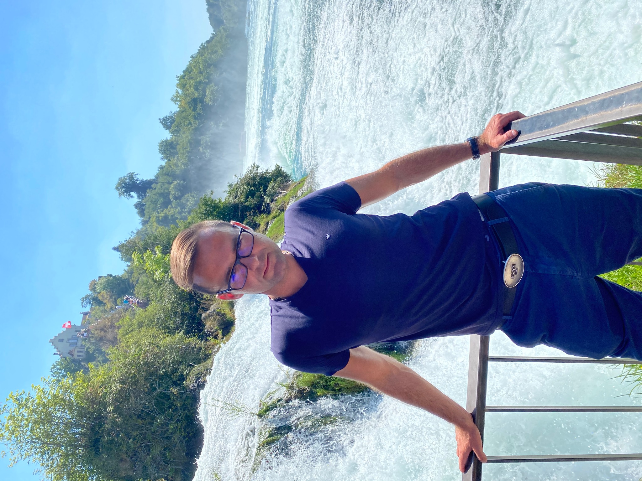 Mężczyzna w okularach, opierający się o metalową barierkę na tle potężnego wodospadu i zieleni drzew, z widocznym zamkiem na wzgórzu w oddali.
