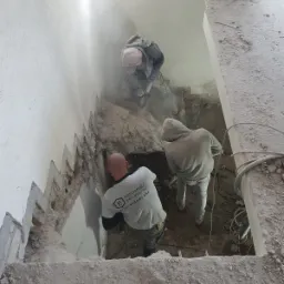 Trzech robotników skuwa starą ścianę schodów, unoszący się pył budowlany, widok z góry.