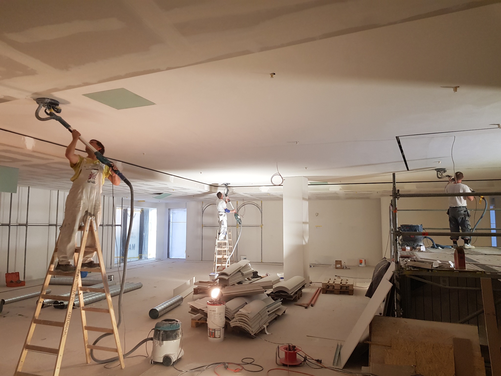 Prace wykończeniowe w dużej przestrzeni: szlifowanie sufitu z płyt kartonowo-gipsowych za pomocą szlifierki z odkurzaczem przez pracownika na drabinie, widoczne rusztowania i materiały budowlane.