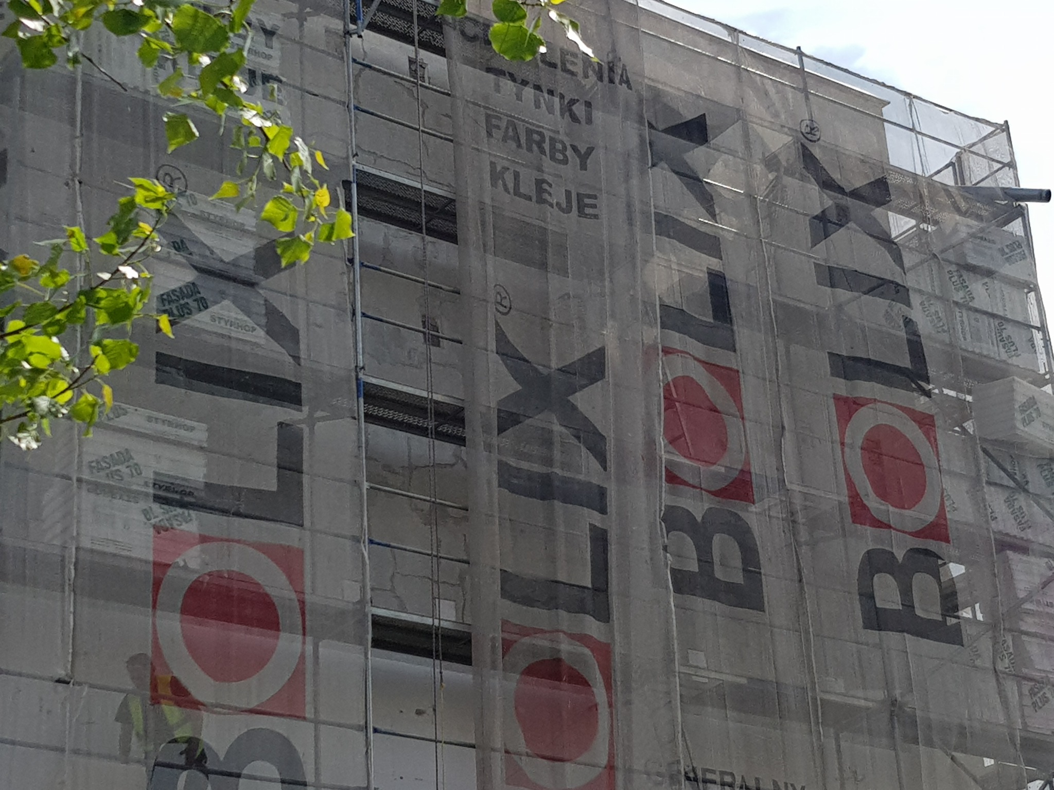 Budynek w trakcie prac elewacyjnych, osłonięty siatką z reklamą materiałów budowlanych, widoczne rusztowania i fragmenty zielonych liści