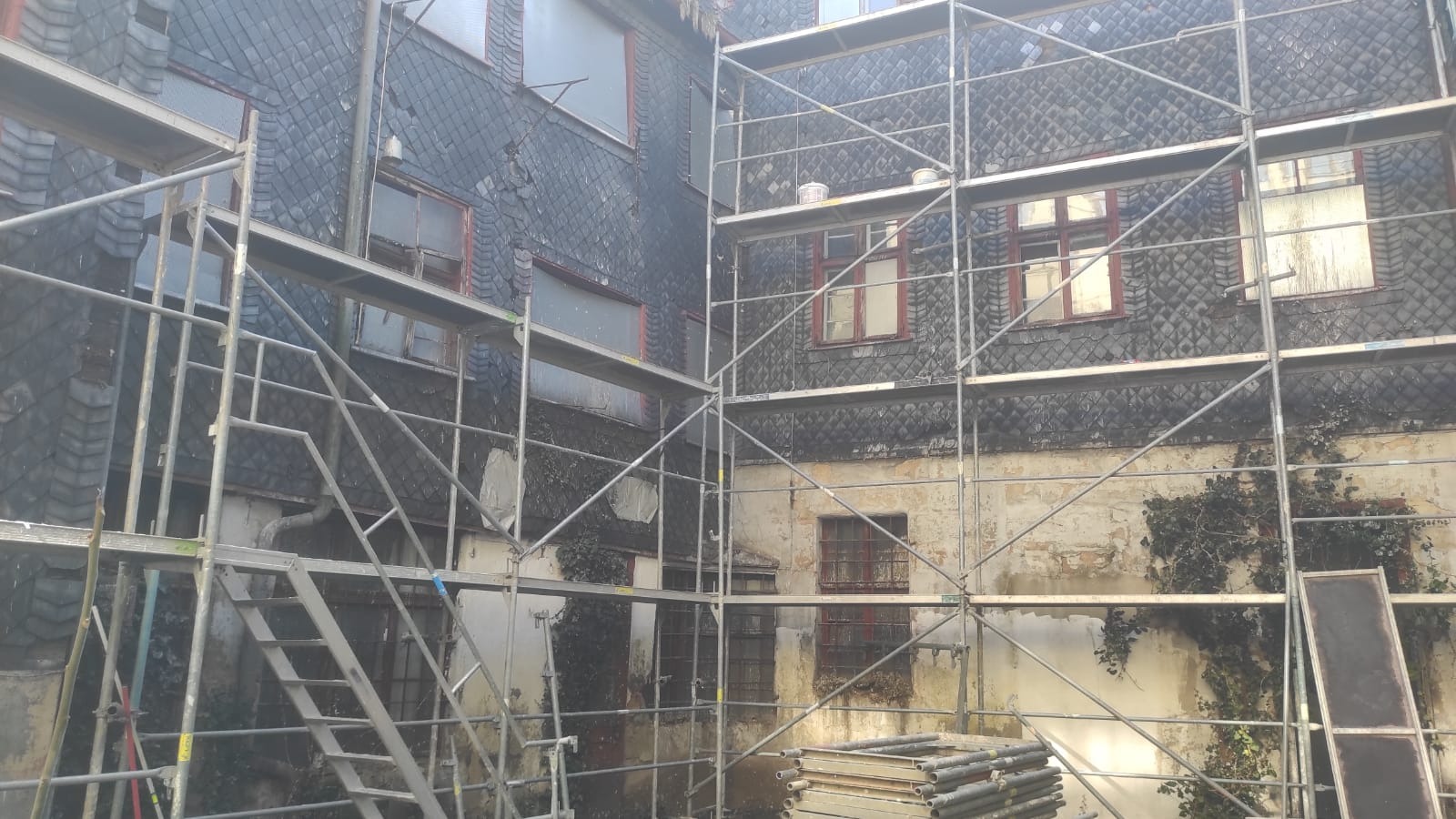 Elewacja budynku z łupka w trakcie renowacji, widoczne rusztowania, fragmenty odpadającego tynku i bluszcz pnący się po ścianie.