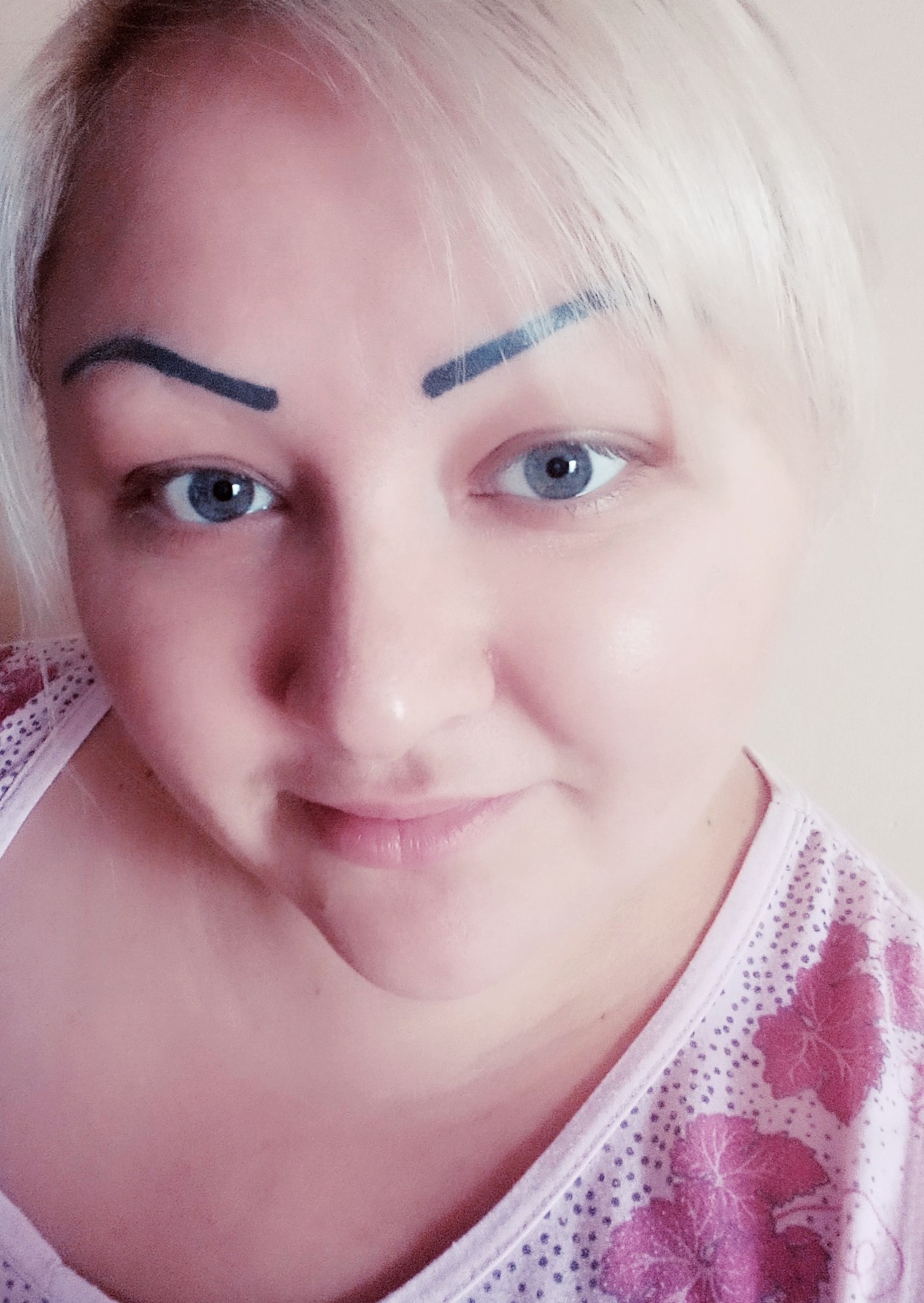 Portret kobiety z blond włosami i wyraźnie podkreślonymi, ciemnymi brwiami, patrzącej w obiektyw z delikatnym uśmiechem.