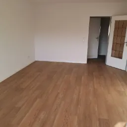 Pusty pokój z drewnianą podłogą z paneli, białe ściany, otwarte drzwi wewnętrzne z szybami w kratkę, wiszące kable od lampy.