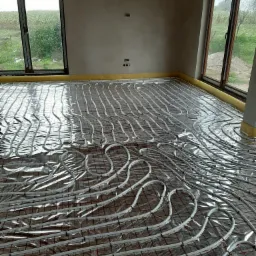 Rozłożona instalacja ogrzewania podłogowego na folii izolacyjnej z widocznymi oknami w surowym stanie wykończenia wnętrza.