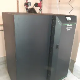 Szary piec na paliwo stałe marki Kostrzewa Twin Bio Luxury Compact z widocznymi rurami miedzianymi i pompami obiegowymi w tle, zainstalowany w pomieszczeniu z jasnymi płytkami.