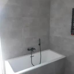 Minimalistyczna łazienka z wanną obudowaną szarymi płytkami, czarną armaturą i grzejnikiem drabinkowym na tle szarych płytek ściennych.