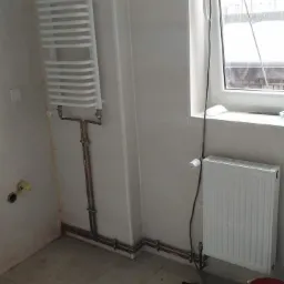 Biała łazienka w trakcie remontu, z nowo zamontowanym grzejnikiem drabinkowym i grzejnikiem panelowym pod oknem, widoczne miedziane rury, narzędzia i wiadro na podłodze.