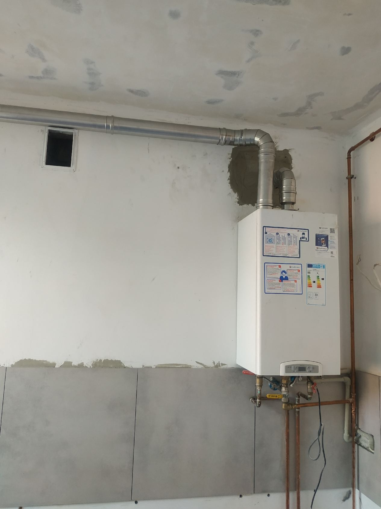 Biały piec grzewczy z podłączonymi rurami, widoczne przyłącza gazowe i wodne, ściana w trakcie wykończenia z częściowo położonymi szarymi płytkami.