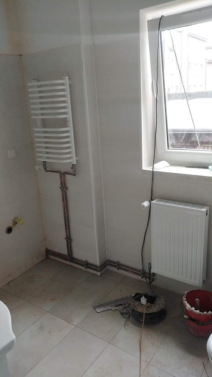 Biała łazienka w trakcie remontu, z nowo zamontowanym grzejnikiem drabinkowym i grzejnikiem panelowym pod oknem, widoczne miedziane rury, narzędzia i wiadro na podłodze.