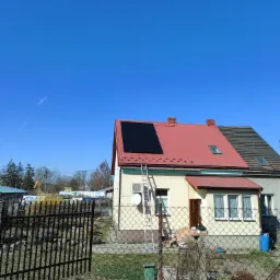 Dwukondygnacyjny dom z czerwonym blaszanym dachem, na którym zamontowano czarne panele słoneczne. Aluminiowa drabina oparta o ścianę. Widoczne ogrodzenie posesji i fragment otoczenia.