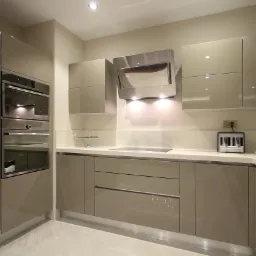 Beżowa, nowoczesna kuchnia z błyszczącymi frontami, zabudowanym piekarnikiem i płytą grzewczą, oświetlona punktowymi lampami sufitowymi, z widocznym ekspresem do kawy i zestawem filiżanek.