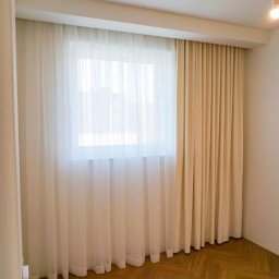 MG-BUD Marcin Głąb - Jasne, przestronne wnętrze w Nowym Sączu z oknem przysłoniętym delikatnymi, białymi firanami i kremowymi zasłonami, uzupełnione parkietem ułożonym we wzór jodełki.