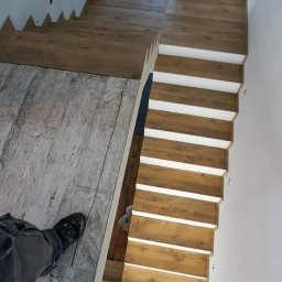 MG-BUD Marcin Głąb - Schody wyłożone panelami drewnopodobnymi z białymi podstopnicami, widok z góry, fragment podłogi z szarych paneli i but pracownika w kadrze.