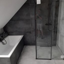 MG-BUD Marcin Głąb - Nowoczesna łazienka na poddaszu z ciemnymi płytkami, białą wanną i kabiną prysznicową z czarnymi ramami; widoczne odbicie fotografa w szybie kabiny.