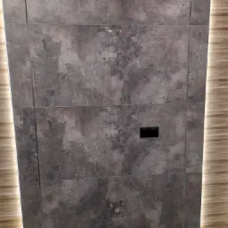 Ściana wyłożona dużymi, grafitowymi płytkami imitującymi beton, z widoczną instalacją wodną i czarnym przyciskiem spłuczki, obok ściana z drewnopodobnych paneli z podświetleniem LED.