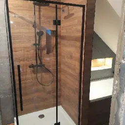 Kabina prysznicowa z czarnymi profilami na tle drewnopodobnych płytek w nowocześnie wykończonej łazience z zabudową w skosie.