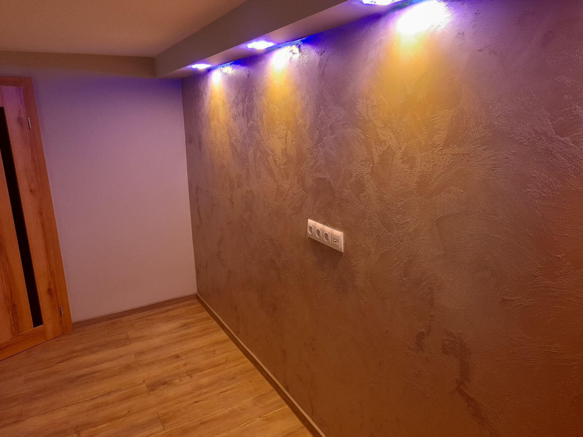 Ściana z dekoracyjną fakturą w odcieniach brązu, oświetlona od góry niebieskimi i żółtymi lampkami LED, z widocznym gniazdkiem elektrycznym i fragmentem drewnianej podłogi.