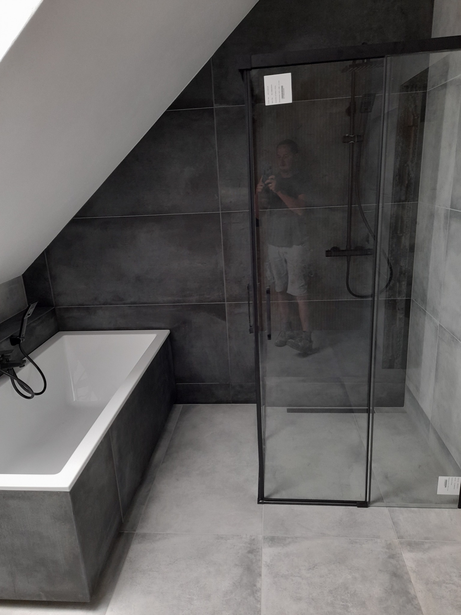 Nowoczesna łazienka na poddaszu z ciemnymi płytkami, białą wanną i kabiną prysznicową z czarnymi ramami; widoczne odbicie fotografa w szybie kabiny.