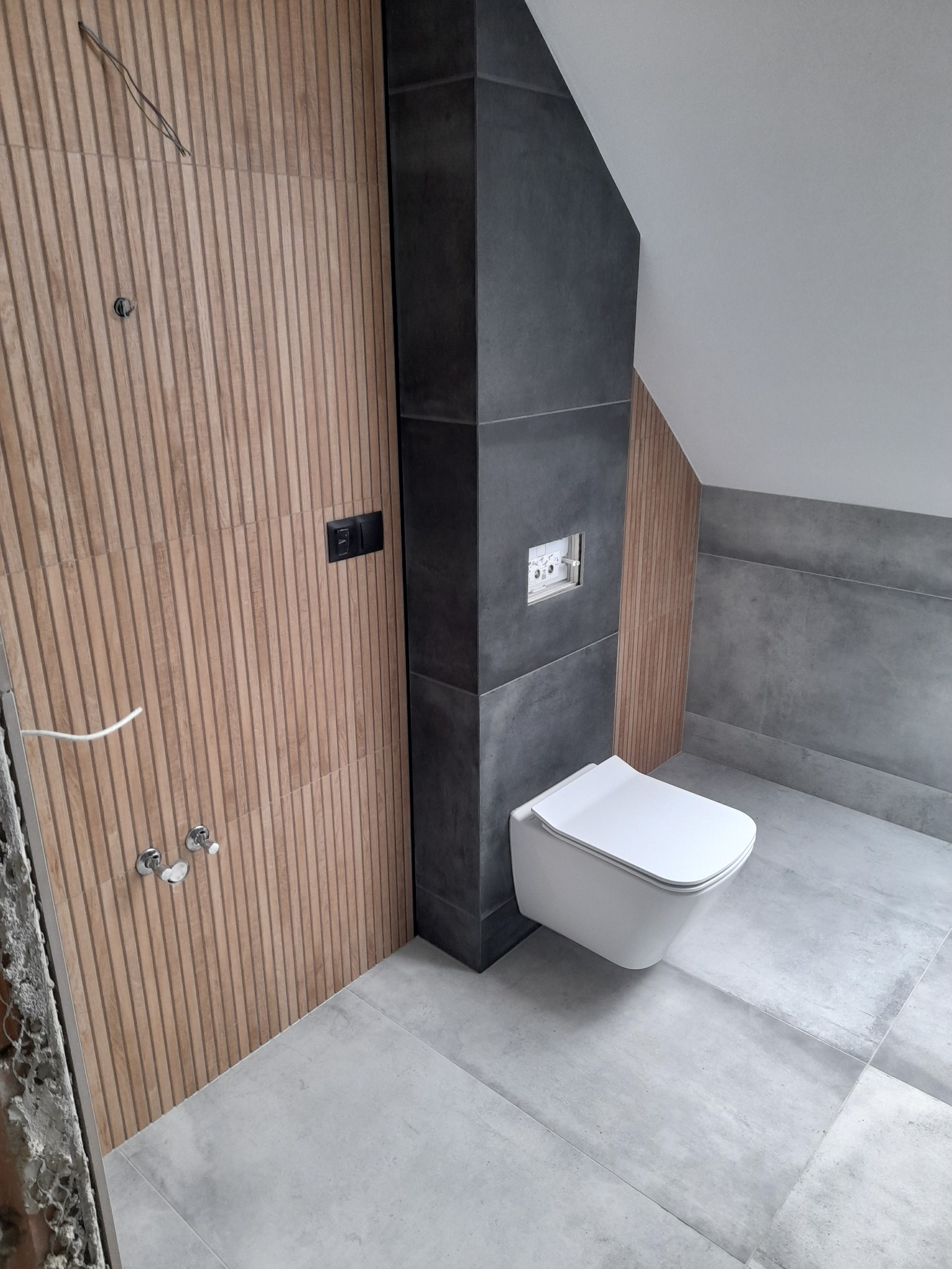 Nowoczesna łazienka w trakcie wykańczania, z wiszącą toaletą, drewnianą okładziną ścienną z pionowymi listwami i szarą płytką podłogową, widoczne wyprowadzenia instalacji elektrycznej i wodnej.