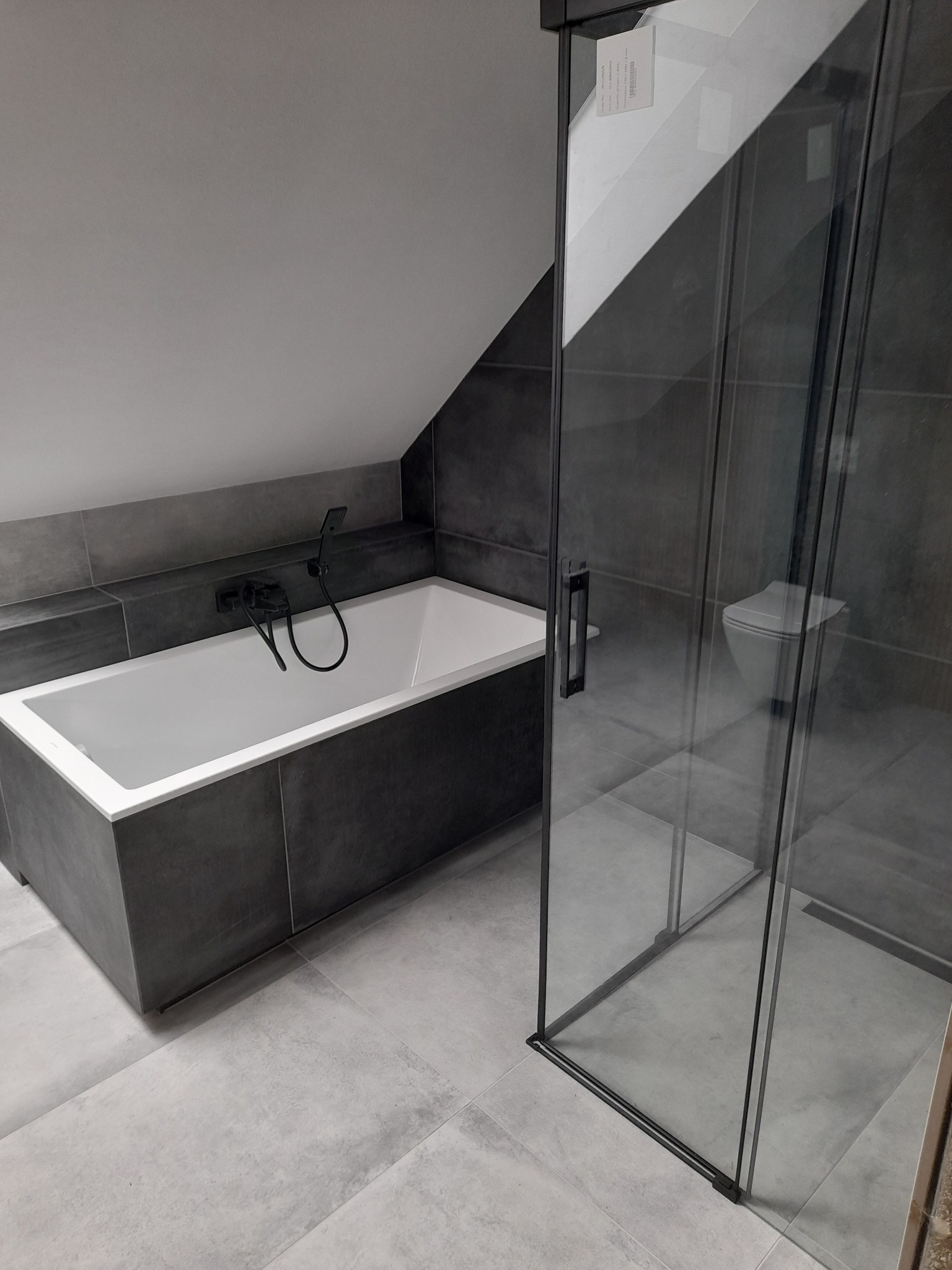 Nowoczesna łazienka na poddaszu z wanną obudowaną szarą płytką, czarną armaturą i kabiną prysznicową ze szklanymi drzwiami z czarnymi ramami, widok z perspektywy podłogi.