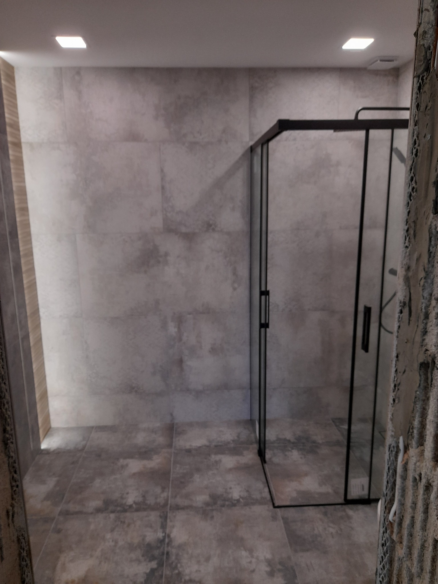 Nowoczesna kabina prysznicowa z czarnymi profilami, szare płytki imitujące beton na ścianach i podłodze, widok z fragmentem surowej ściany w tle.