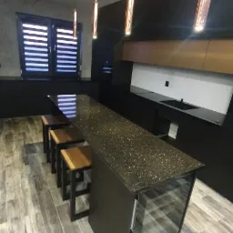 Nowoczesna kuchnia z czarnymi szafkami, blatem z połyskującymi drobinkami i drewnianymi hokerami, oświetlona wiszącymi lampami w kształcie tub w kolorze miedzi.