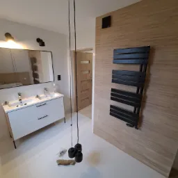 Nowoczesna łazienka z białymi płytkami imitującymi marmur, podwójna umywalka z szafką w kolorze białym z czarnymi uchwytami, czarny grzejnik drabinkowy na ścianie z drewnopodobnych paneli...