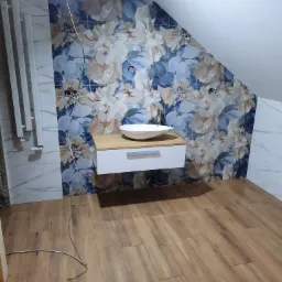 Ukończona łazienka na poddaszu z drewnopodobną podłogą, umywalką nablatową na białej szafce i ścianą w kwiatowy wzór z płytek ceramicznych, grzejnik drabinkowy po lewej.