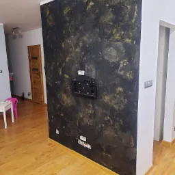 Ściana akcentowa w pokoju, pomalowana na czarno ze złotymi smugami, z zamontowanym uchwytem na telewizor i widocznymi gniazdkami elektrycznymi, na podłodze panele w odcieniach brązu.