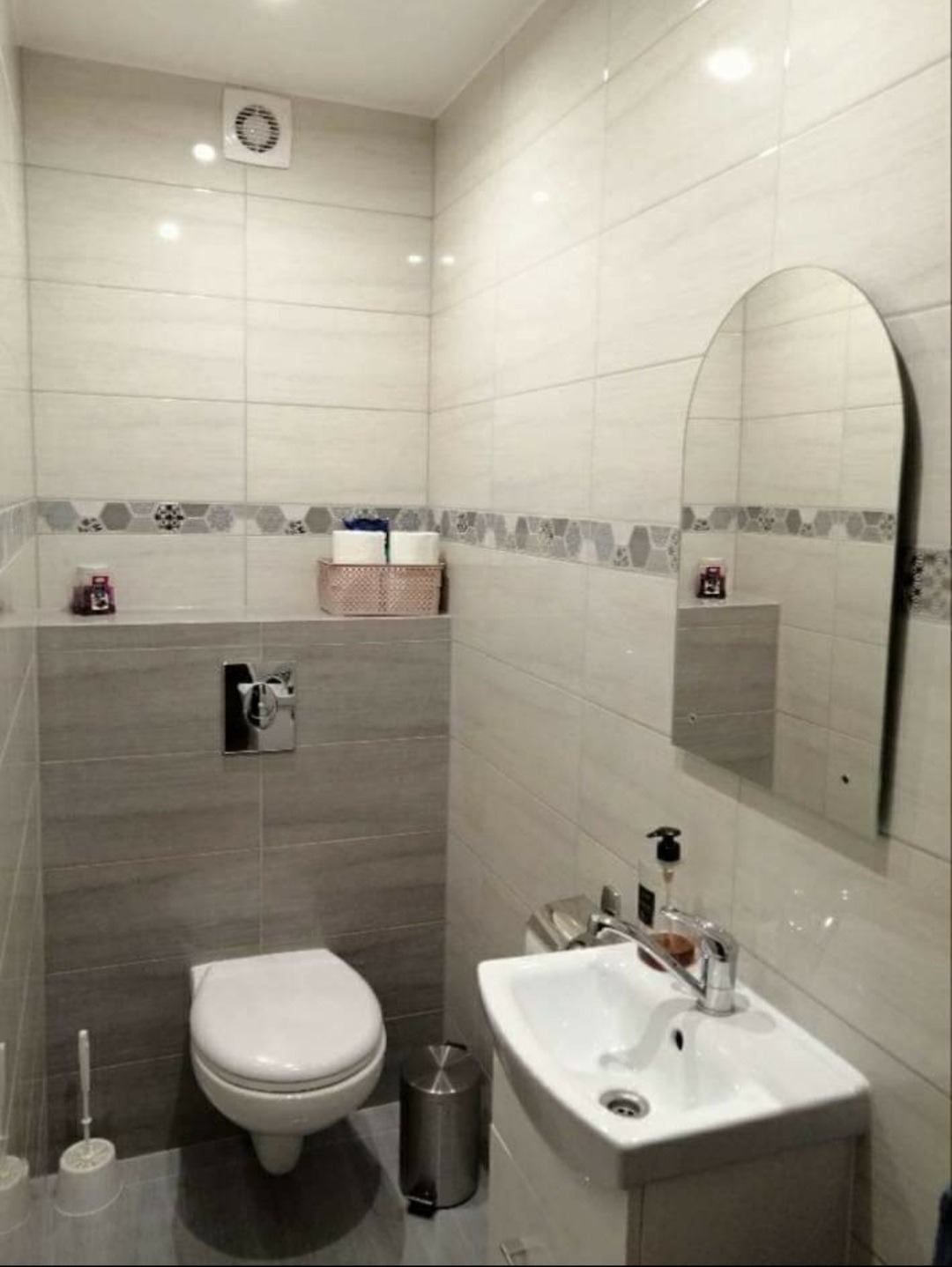 Wykończona łazienka z toaletą podwieszaną, umywalką z szafką i dużym lustrem w zaokrąglonej ramie, wyłożona płytkami w odcieniach szarości i bieli z dekoracyjnym pasem mozaiki.