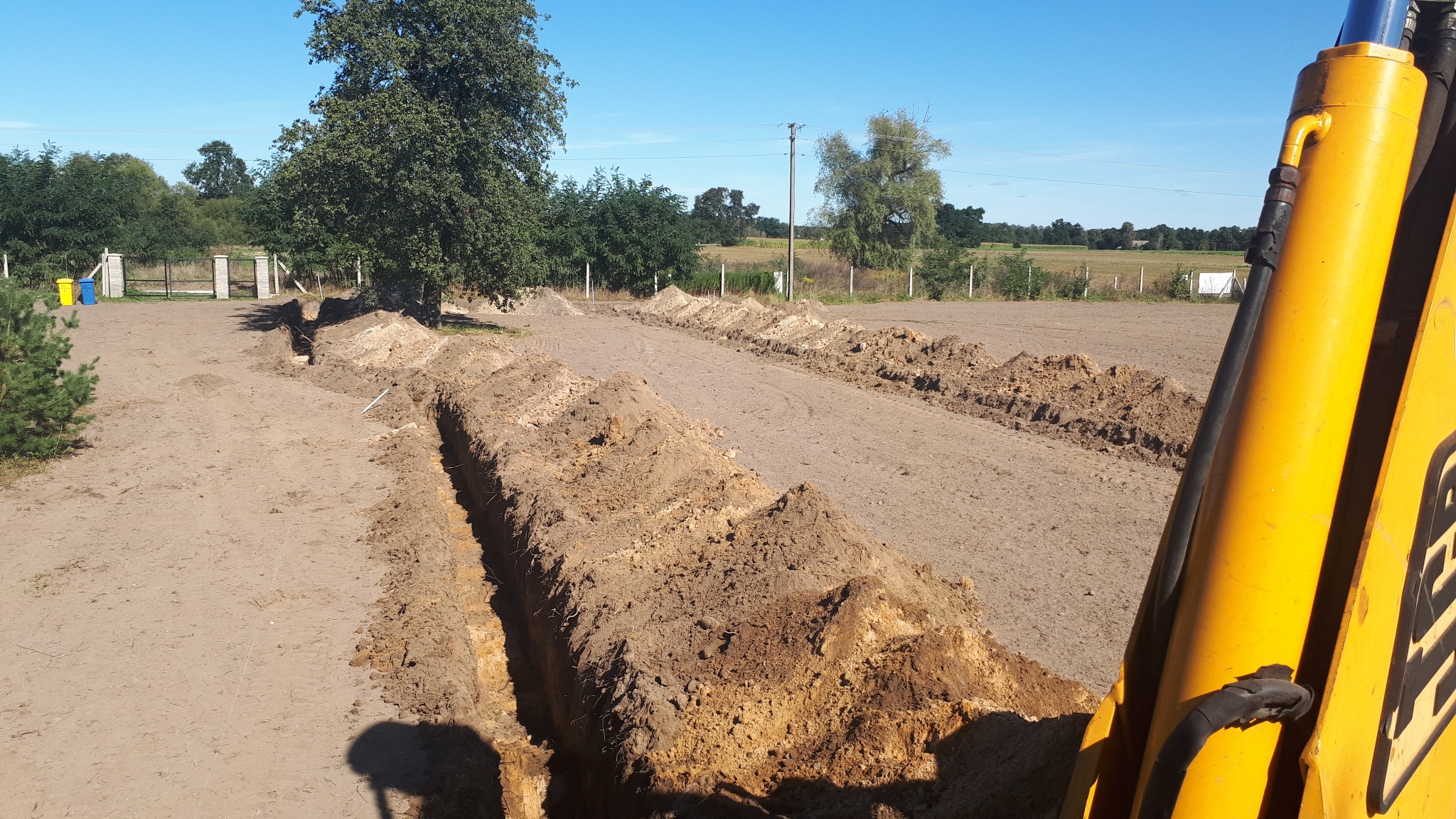 Wykopane rowy w ziemi pod system nawadniania, widoczny fragment koparki, w tle drzewa i błękitne niebo.