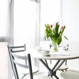 Jasne wnętrze mieszkania z okrągłym stołem, bukietem tulipanów w wazonie, filiżankami i szarymi krzesłami, widok na budynek przez okno.