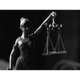 Czarno-biała statuetka Temidy z zasłoniętymi oczami, trzymająca w górze wagę sprawiedliwości na tle ciemnego tła.