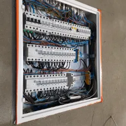 Otwarta skrzynka elektryczna z zainstalowanymi bezpiecznikami EATON, widoczne liczne kable w kolorach niebieskim, brązowym i czarnym, montaż w surowej ścianie z widoczną siatką.