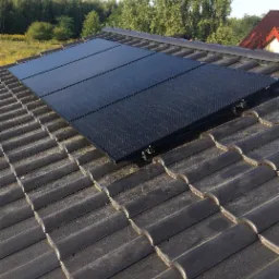 Cztery czarne panele fotowoltaiczne zamontowane na ciemnoszarym dachu z dachówki ceramicznej, widok z góry na tle zieleni drzew i błękitnego nieba.