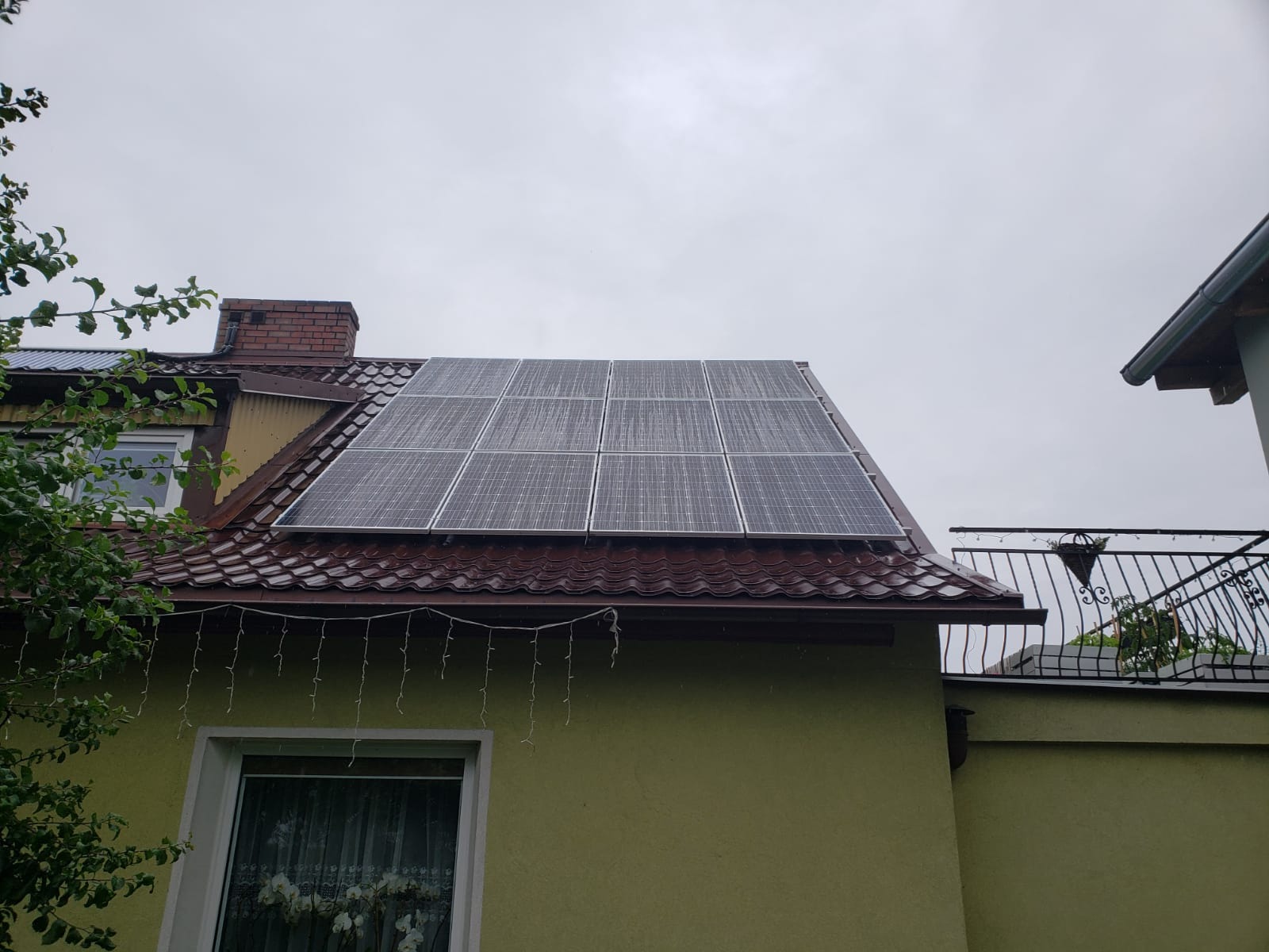 Panele słoneczne na dachu domu z czerwoną dachówką, widoczne z boku w pochmurny dzień.