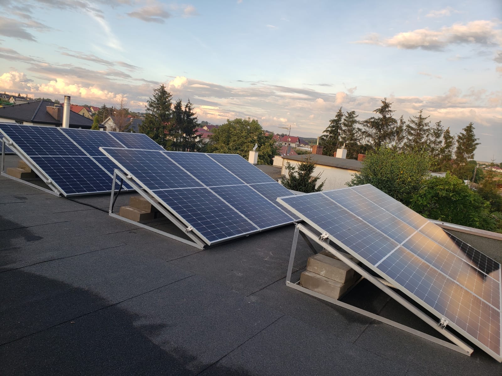 Panele słoneczne na dachu z widokiem na zabudowania w Koninie, montowane na metalowej konstrukcji opartej na betonowych bloczkach, w tle błękitne niebo z chmurami.