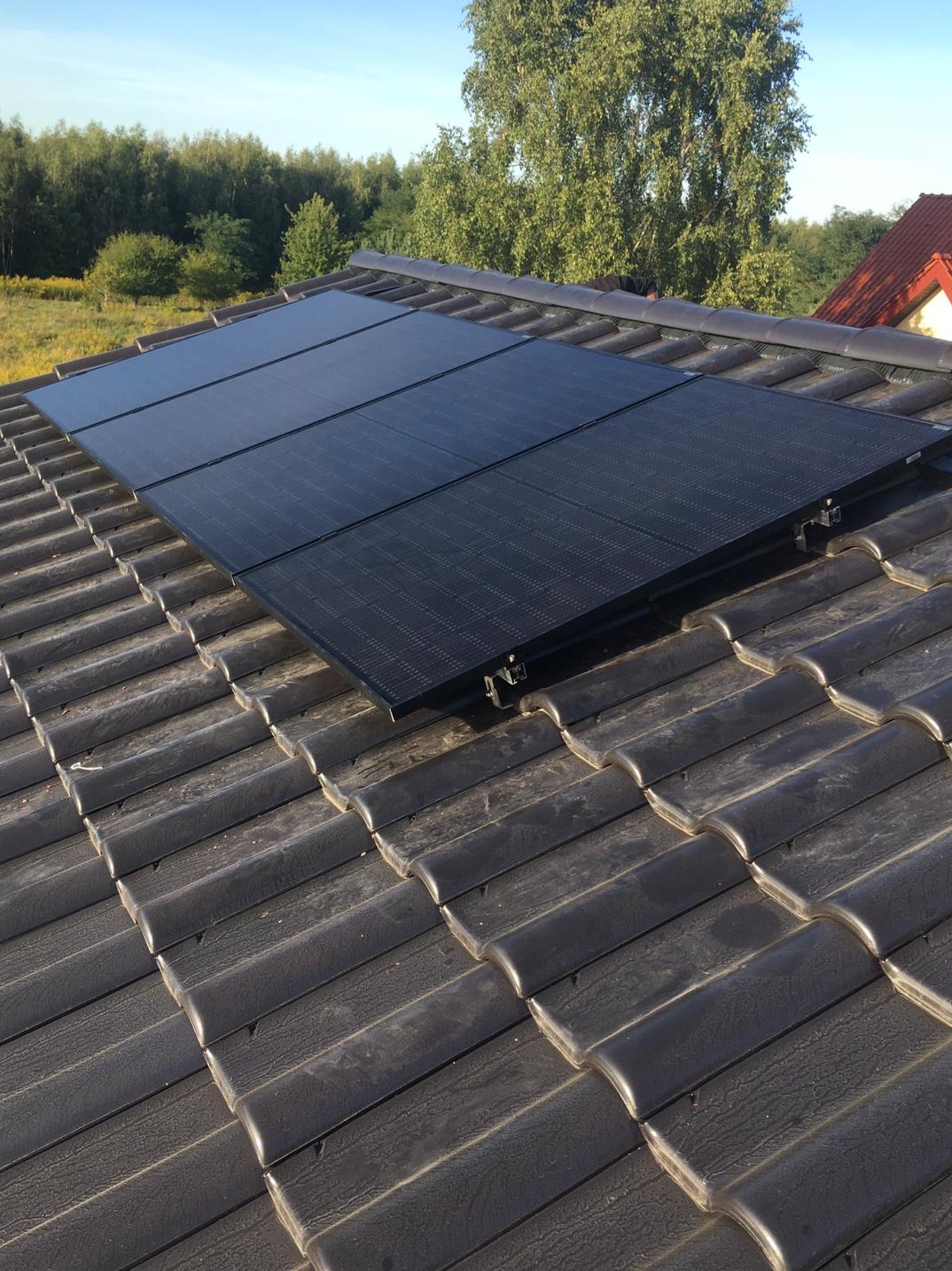 Cztery czarne panele fotowoltaiczne zamontowane na ciemnoszarym dachu z dachówki ceramicznej, widok z góry na tle zieleni drzew i błękitnego nieba.