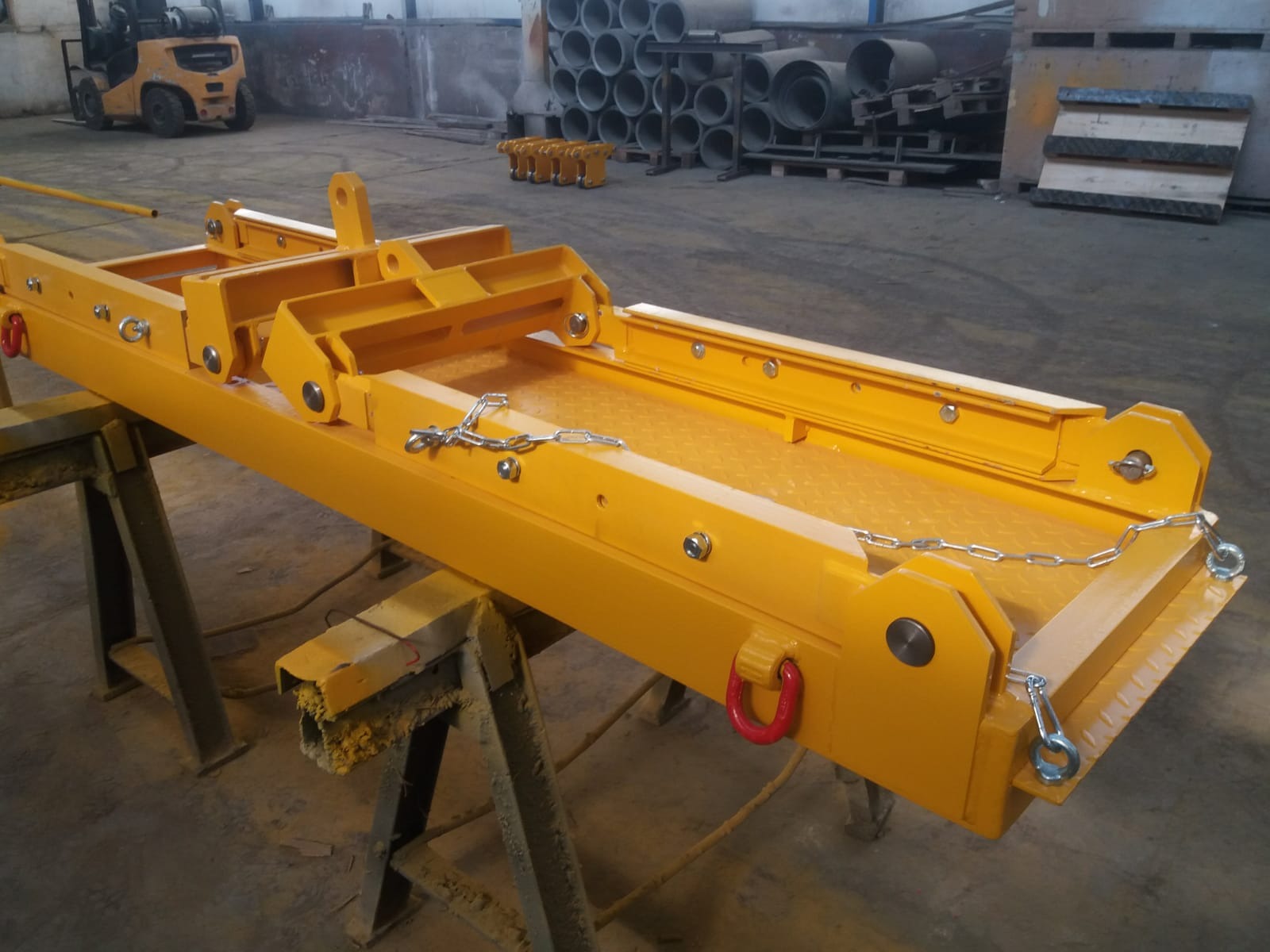 Żółty, stalowy uchwyt transportowy na kozłach warsztatowych z widocznym w tle wózkiem widłowym i stosem rur.