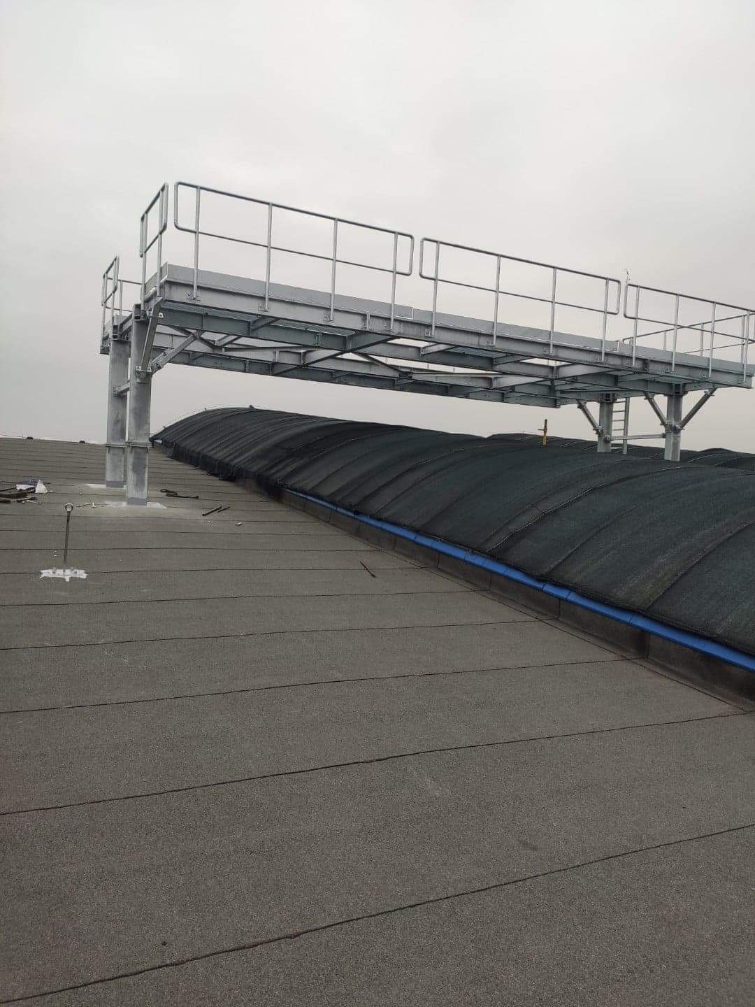 Metalowa platforma serwisowa z barierkami, zamontowana na płaskim dachu pokrytym papą, z widocznym fragmentem dachu hali w tle.