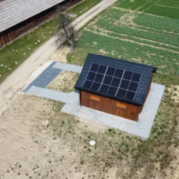 Widok z drona na mały drewniany budynek z panelami słonecznymi na dachu, otoczony polami uprawnymi i zabudowaniami gospodarczymi.