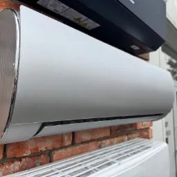 Srebrny klimatyzator ścienny na tle ceglanej ściany, widoczne inne jednostki klimatyzacyjne powyżej i poniżej, obok przeszklone drzwi.