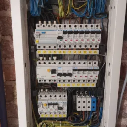 Otwarta skrzynka rozdzielcza z licznymi bezpiecznikami, przewodami i złączkami, zamontowana na ścianie z cegły.