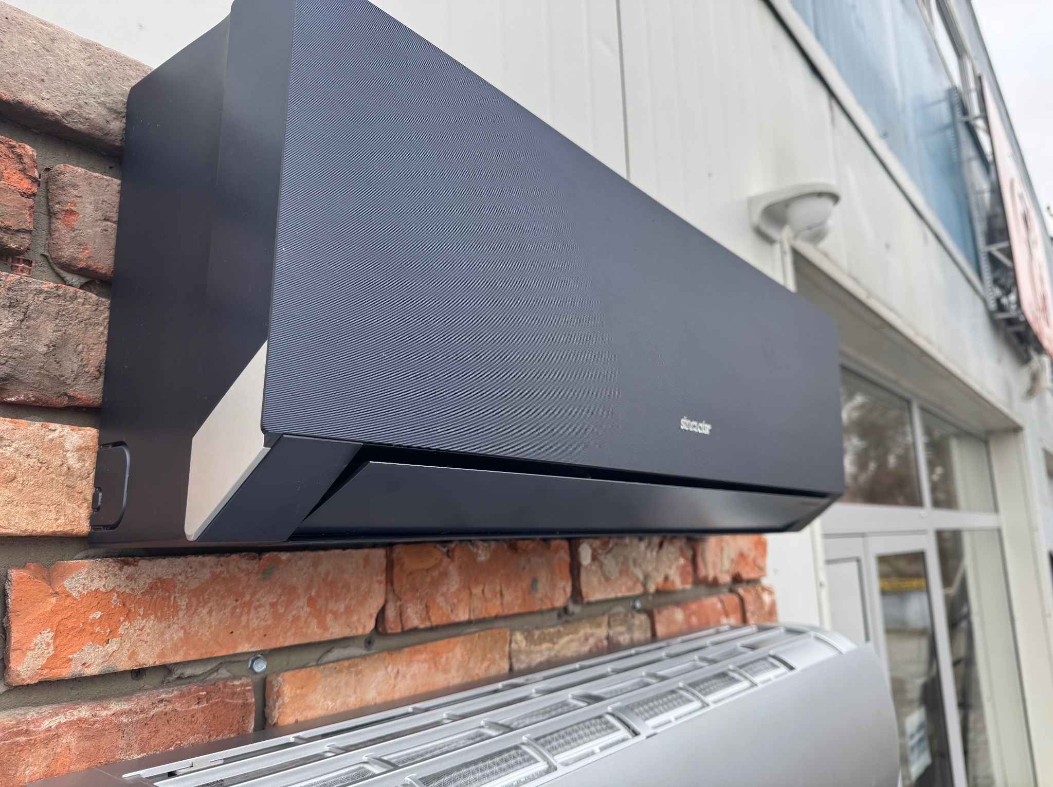 Czarny klimatyzator ścienny zamontowany na ceglanej ścianie, widoczny fragment drugiego klimatyzatora poniżej.